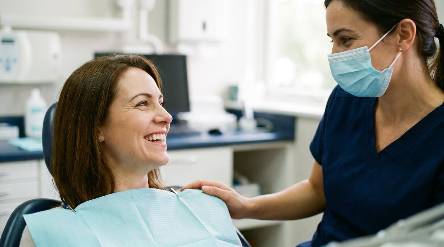 A patient in a dental chair smiling while a clinician in scrubs and mask rests a reassuring hand on her shoulder